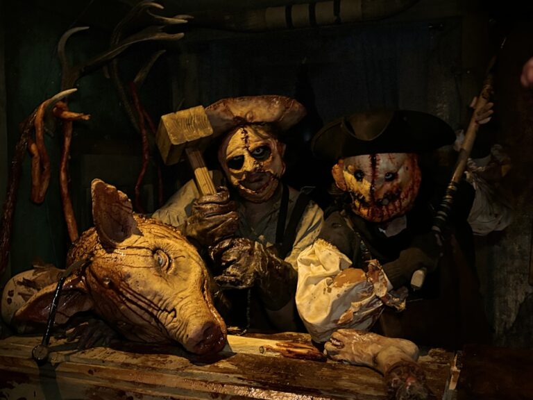 Two people in gruesome costumes pose behind a bloody butchered pig's head, holding a hammer and a knife, in a dimly lit, horror-themed setting.